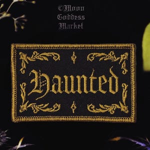 May include: A rectangular black embroidered patch featuring the word Haunted in gold gothic lettering. The text is surrounded by an ornate gold border with decorative flourishes. Above the patch, the text Moon Goddess Market is visible.