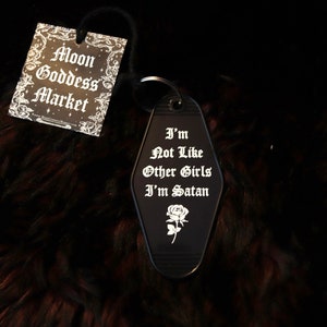 May include: Black motel key chain with the text "I'm Not Like Other Girls I'm Satan" and a rose design. The keychain is on a brown fuzzy surface.