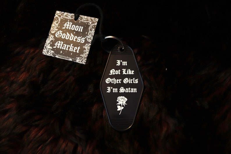 May include: Black motel key chain with the text "I'm Not Like Other Girls I'm Satan" and a rose design. The keychain is on a brown fuzzy surface.