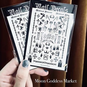 May include: Two packs of nail decals featuring medieval-themed designs. Each pack displays black and white illustrations of dragons, crosses, keys, and other medieval elements. The text "Nail Decals" and "Medieval Princess" are visible on the packaging. The brand name "Moon Goddess Market" is also displayed.