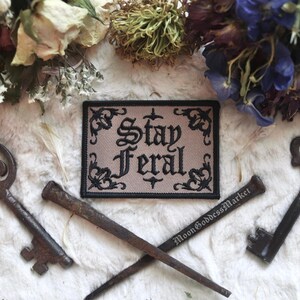 May include: Black and white embroidered patch with the text "Stay Feral" on a beige background. The patch is surrounded by a black border with decorative floral designs.