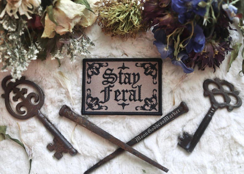 May include: Black and white embroidered patch with the text "Stay Feral" on a beige background. The patch is surrounded by a black border with decorative floral designs.