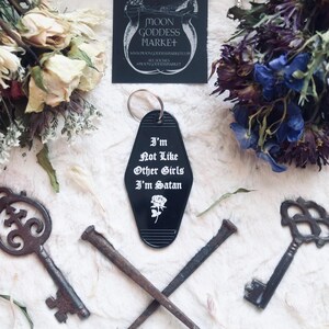 May include: Black motel key chain with white text that reads "I'm Not Like Other Girls I'm Satan" with a white rose graphic.