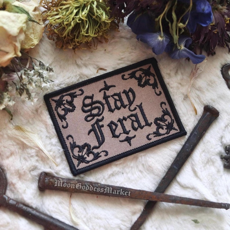May include: A beige fabric patch with black embroidered lettering that reads "Stay Feral". The lettering is in a gothic font and is surrounded by a decorative black border.