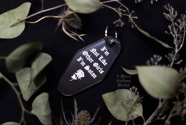 May include: Black metal keychain shaped like a motel key with the text "I'm not like other girls I'm Satan" in white gothic font and a small white rose graphic. The keychain is on a black background with green leaves.