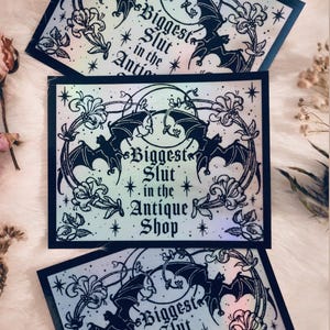 May include: Three rectangular stickers with a black border and a design featuring bats, flowers, and the text "Biggest Slut in the Antique Shop." The stickers have a holographic, iridescent finish.