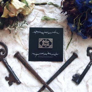 May include: A black and white enamel pin with a gold border and the text "Stay Feral" in a gothic font. The pin is on a black background with a barbed wire design and the text "MoonGoddessMarket".