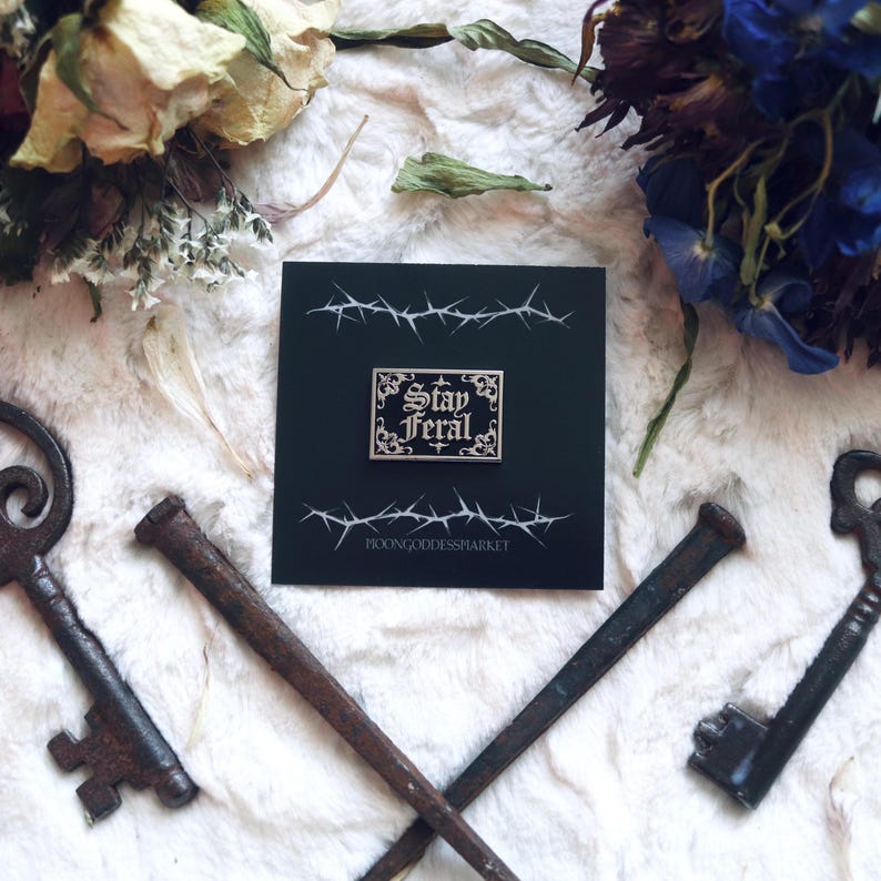 May include: A black and white enamel pin with a gold border and the text "Stay Feral" in a gothic font. The pin is on a black background with a barbed wire design and the text "MoonGoddessMarket".