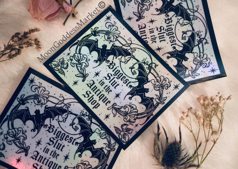 May include: Several square stickers with a holographic finish are displayed. Each sticker features a black bat design, floral accents, and the text "Biggest Slut in the Antique Shop." The stickers are arranged on a soft, white surface with dried flowers.