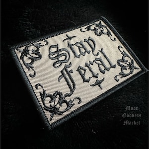 May include: A beige embroidered patch with black thread. The patch features the words "Stay Evil" in a gothic font.