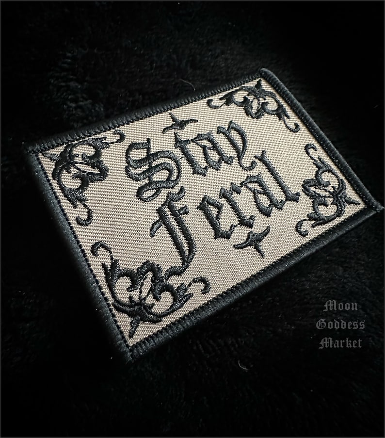 May include: A beige embroidered patch with black thread. The patch features the words "Stay Evil" in a gothic font.