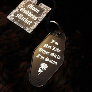 May include: Black metal keychain shaped like a motel key with the text "I'm Not Like Other Girls I'm Satan" and a small white rose. The keychain is on a black fuzzy surface with a tag that says "Moon Goddess Market".