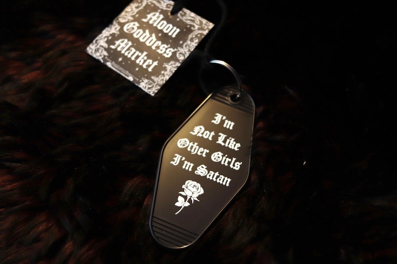 May include: Black metal keychain shaped like a motel key with the text "I'm Not Like Other Girls I'm Satan" and a small white rose. The keychain is on a black fuzzy surface with a tag that says "Moon Goddess Market".