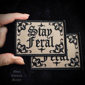May include: Two beige embroidered patches with black stitching. The patches feature a decorative border and the text "Stay Feral" in a gothic font.