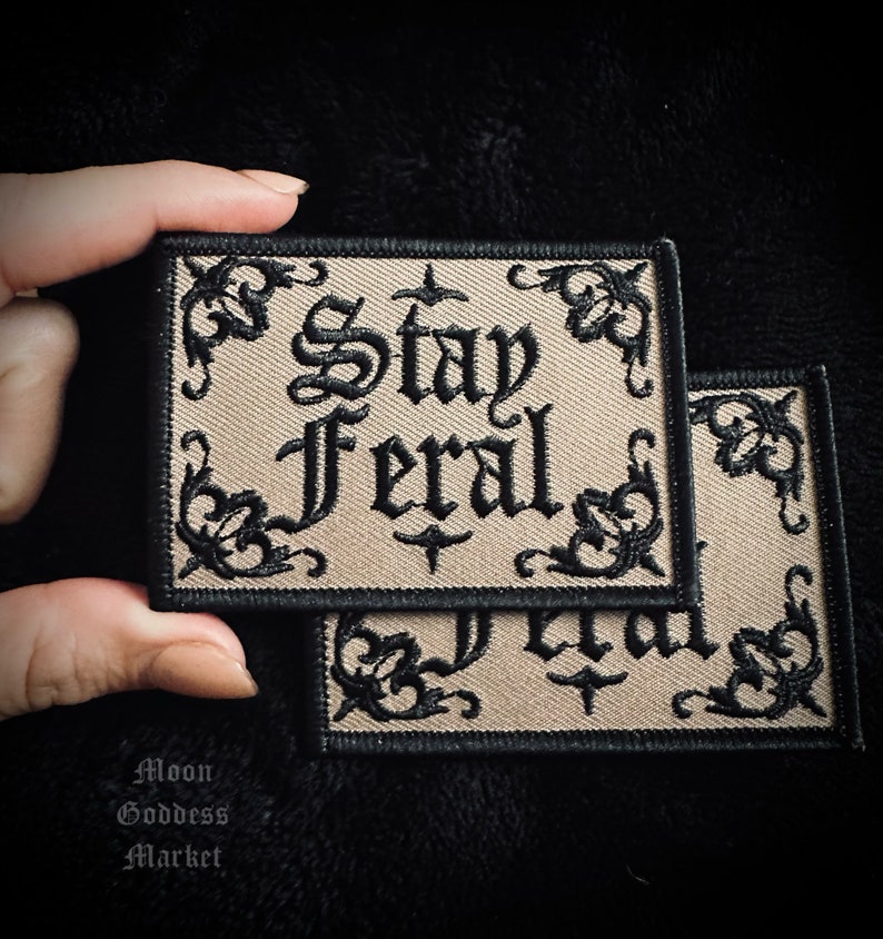 May include: Two beige embroidered patches with black stitching. The patches feature a decorative border and the text "Stay Feral" in a gothic font.