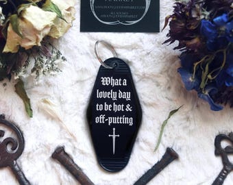 Puede incluir: Un llavero negro estilo motel con un anillo de metal plateado. El llavero presenta texto blanco que dice "What a lovely day to be hot & off-putting" sobre un pequeño icono de espada blanca. El diseño está centrado en una forma de diamante.