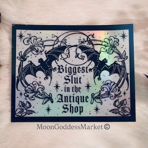 May include: A rectangular sticker with a black border and a holographic background. The sticker features a design with bats, flowers, and the text "Biggest Slut in the Antique Shop." The MoonGoddessMarket logo is at the bottom.