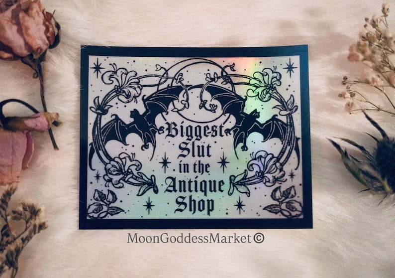 May include: A rectangular sticker with a black border and a holographic background. The sticker features a design with bats, flowers, and the text "Biggest Slut in the Antique Shop." The MoonGoddessMarket logo is at the bottom.