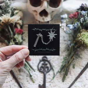 Pode incluir: Um broche prateado com um chicote de pontas com uma corrente. O broche é exibido em um cartão preto com uma borda de arame farpado e o texto "ROCKING JEWELRY MARKET".