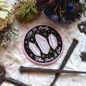 以下が含まれることがあります： 黒い背景に「Life is Rocky When You're a Gem」という文字と、3つのピンクのクリスタル形の刺繍が施されたピンクと黒の刺繍パッチです。