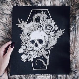 Puede incluir: Impresión artística en lienzo negro con un diseño de calavera blanca, serpiente y flores dentro de un marco en forma de ataúd. La obra de arte incluye el texto "MoonGoddessMarket" en la parte inferior. Tema gótico u oculto.