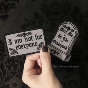 May include: Two iron-on patches with a gothic aesthetic. One patch is a tombstone with the text "Meet Me In The Graveyard". The other patch is rectangular with the text "I am not for everyone".