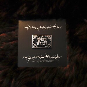 May include: A black and silver enamel pin with the text "Stay Feral" in a gothic font. The pin is on a black background with a barbed wire design.