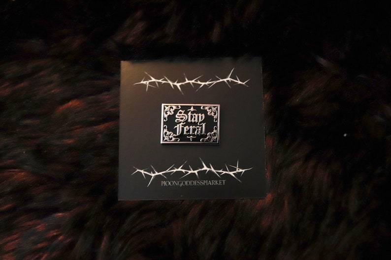 May include: A black and silver enamel pin with the text "Stay Feral" in a gothic font. The pin is on a black background with a barbed wire design.