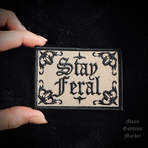 May include: Beige embroidered patch with black stitching and the text "Stay Feral" in a gothic font.