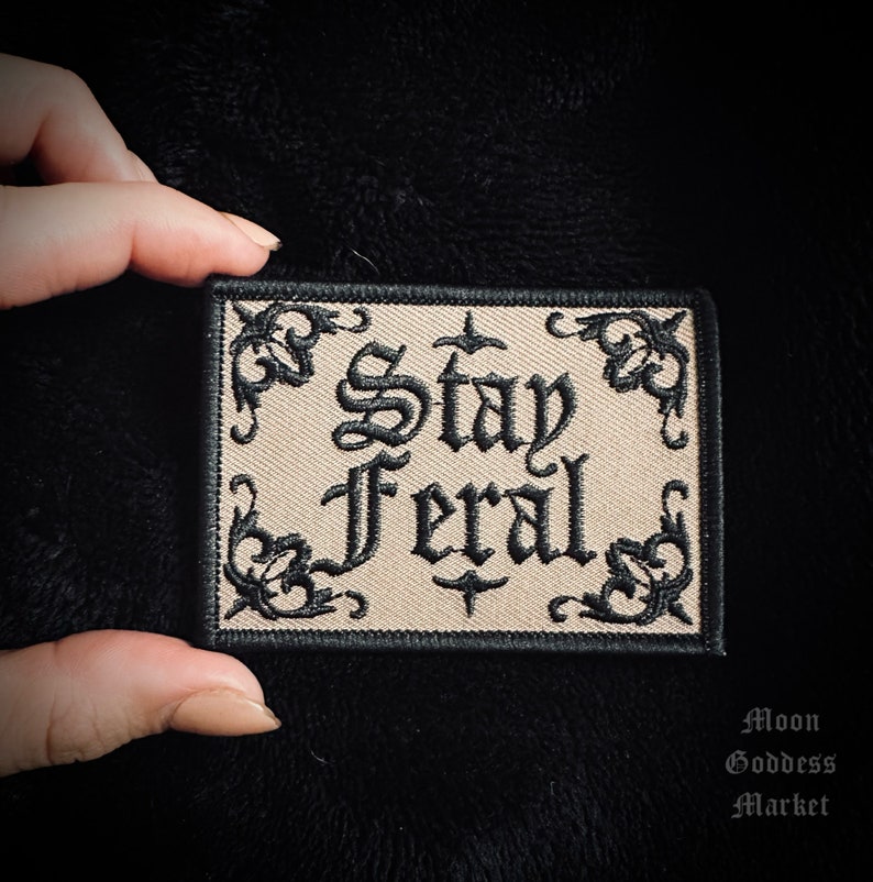 May include: Beige embroidered patch with black stitching and the text "Stay Feral" in a gothic font.