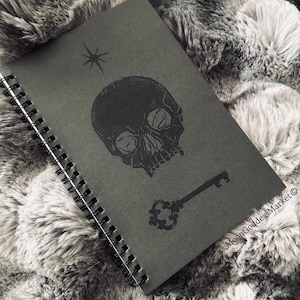 May include: A black spiral-bound notebook with a skull, key, and star design. The notebook's cover features a detailed illustration of a human skull, a vintage key, and a star. The design is printed in black on a dark gray background.