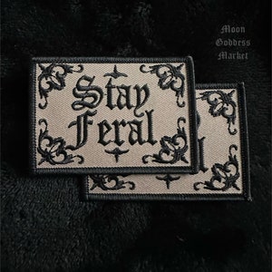May include: Two beige embroidered patches with black stitching. The patches have a decorative border and the text "Stay Feral" in a gothic font.