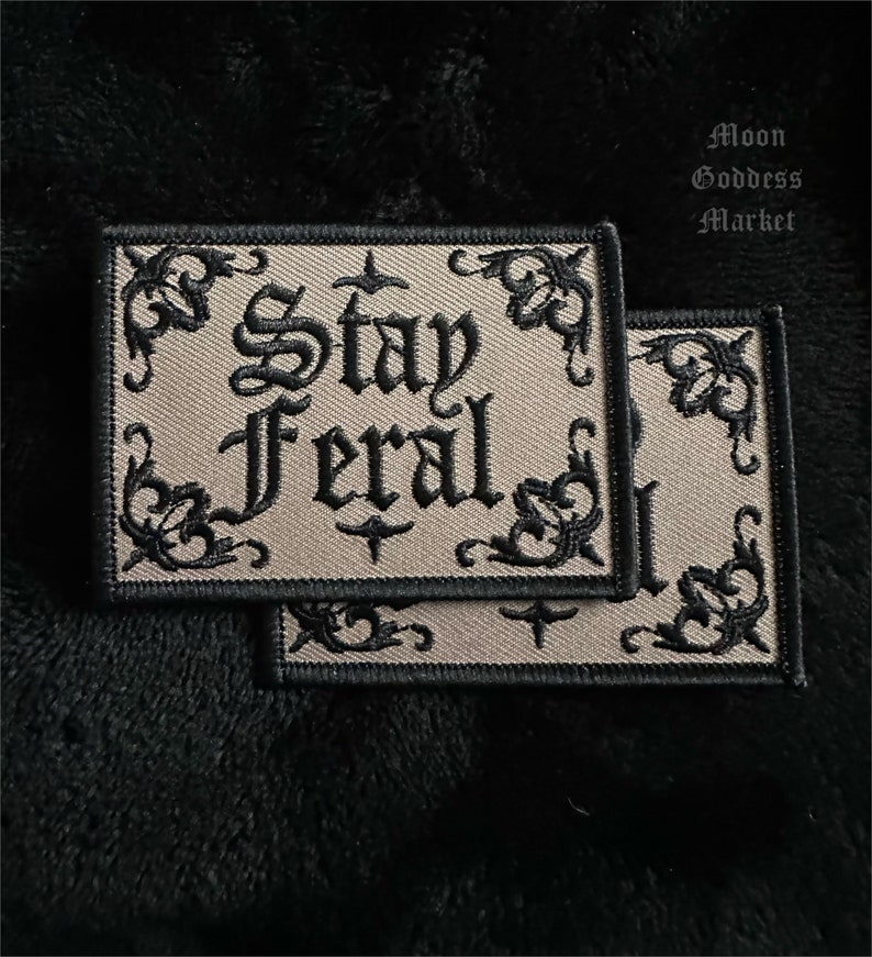 May include: Two beige embroidered patches with black stitching. The patches have a decorative border and the text "Stay Feral" in a gothic font.