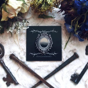 May include: Silver-toned pin with a mirror in the center and the text "Never forget you are more than enough" around the mirror. The pin is on a black background with a thorny vine design.