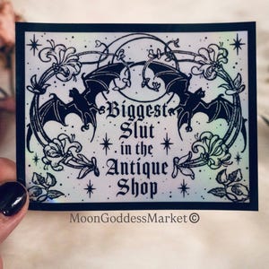 May include: A black and white sticker with the text "Biggest Slut in the Antique Shop". The sticker features two bats, floral accents, and the MoonGoddessMarket logo. The design has a holographic, iridescent effect.