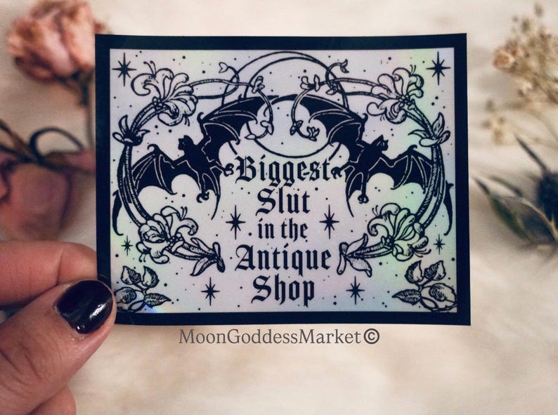 May include: A black and white sticker with the text "Biggest Slut in the Antique Shop". The sticker features two bats, floral accents, and the MoonGoddessMarket logo. The design has a holographic, iridescent effect.