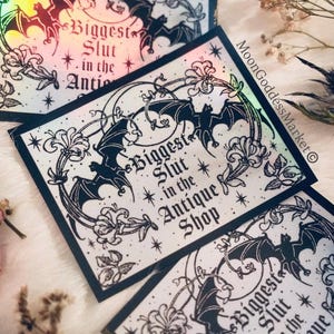 May include: Holographic stickers with a black bat and floral design, featuring the text "Biggest Slut in the Antique Shop." The stickers have a rainbow sheen and are surrounded by dried flowers.