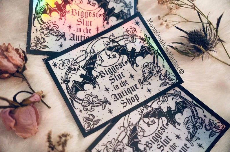 May include: Holographic stickers with a black bat and floral design, featuring the text "Biggest Slut in the Antique Shop." The stickers have a rainbow sheen and are surrounded by dried flowers.