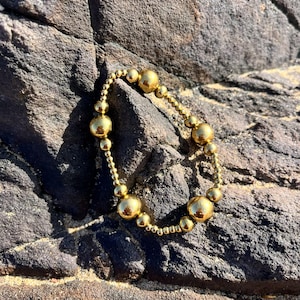 Puede incluir: Una pulsera circular compuesta por cuentas esféricas de color dorado pulido de diferentes tamaños. Las cuentas están dispuestas en un patrón repetitivo, alternando entre esferas metálicas más pequeñas y más grandes, creando una superficie reflectante.