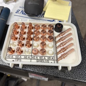 May include: A white plastic organizer tray containing copper plasma cutter consumables, including various tips, shields, and electrodes. The tray is labeled with text indicating 40 A, 60 A, and 80 A ratings for the tips and shields, organized in rows.