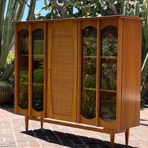May include: A tall wooden cabinet featuring two glass-paneled doors on the sides and a central door with a woven cane panel. The piece stands on four tapered legs and includes internal shelving visible through the glass sections of the frame.