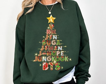 Può includere: Una felpa girocollo verde bosco con la grafica di un albero di Natale composto da lettere in stile pan di zenzero. L'albero è sormontato da una stella dorata e riporta i nomi RM, JIN, SUGA, JIMIN, J-HOPE, JUNGKOOK e BTS.