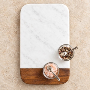 May include: A rectangular serving board featuring a white marble top section with grey veining and a dark brown wood bottom section. Two small glass bowls sit on the board, one containing pink salt and the other mixed peppercorns, each with a silver spoon.