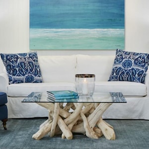 May include: A coffee table with a clear glass rectangular top supported by a base made of light-colored, intertwined driftwood branches. The table is positioned in front of a white sofa with two blue patterned throw pillows and a blue painting.