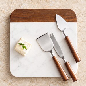May include: A square serving board with a white marble base and a dark wood top edge. Three stainless steel cheese knives with matching dark wood handles are arranged on the board next to a small square piece of white cheese with a green herb.