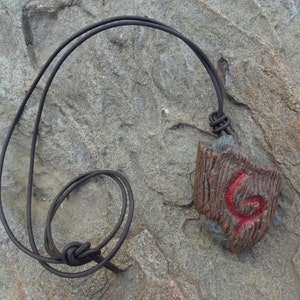 Puede incluir: Un collar con colgante marrón con un cordón de cuero. El colgante tiene una textura de madera y un diseño de espiral roja. El collar se muestra sobre una superficie de piedra gris.