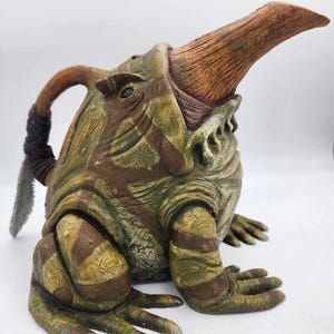 May include: A ceramic frog sculpture with a long, pointed snout and a textured, green and brown patterned body. The frog has a wide open mouth and a large, round eye.