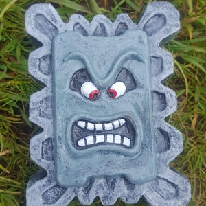 May include: A gray concrete garden statue with a face. The face has red eyes, white teeth, and a frowning expression. The statue is surrounded by green grass.