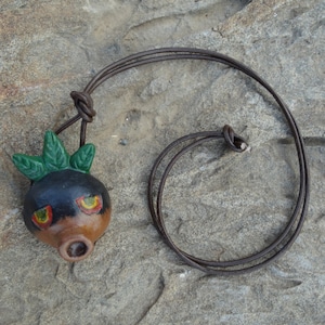 Puede incluir: Un collar de cordón de cuero marrón con un colgante de cerámica. El colgante está pintado para parecer una cara con hojas verdes en la parte superior. La cara tiene cabello negro, ojos rojos y una nariz marrón.