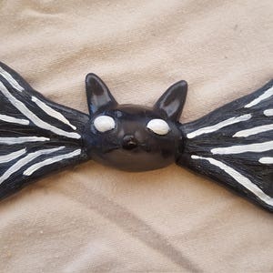 May include: A black bat-shaped figurine with white stripes on the wings and white eyes. The bat has a cat-like face and is made of a dark, possibly painted material. The wings are spread out.